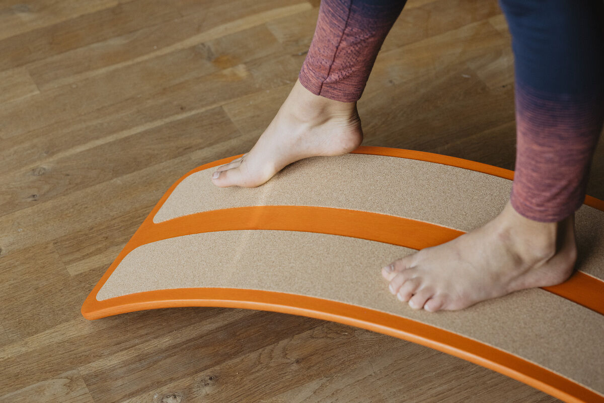 Nahaufnahme von nackten Füßen auf einem Balance Board »das.Brett« in pompejanisch rot, stehend auf einem Parkettboden. Das Balance Board zeigt eine leichte Wölbung und hat beige und rote Bereiche.