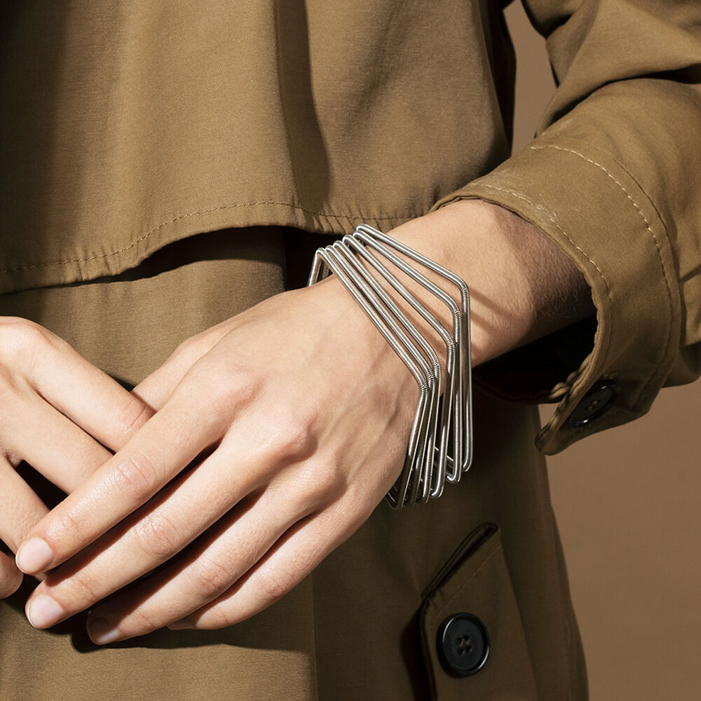 Ein silbernes Armband mit quadratischen Metallsegmenten ist am Handgelenk einer Person zu sehen, die eine beigefarbene Jacke trägt.