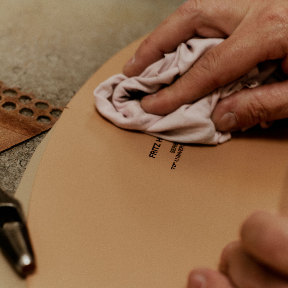 Nahaufnahme einer Hand, die mit einem Tuch die Sitzfläche eines beigen Stuhls vom Modell »Series 7™ Tailored« der Marke Fritz Hansen poliert. Einige Werkzeuge und ein Stück gestanztes Leder liegen daneben auf einer Werkbank.
