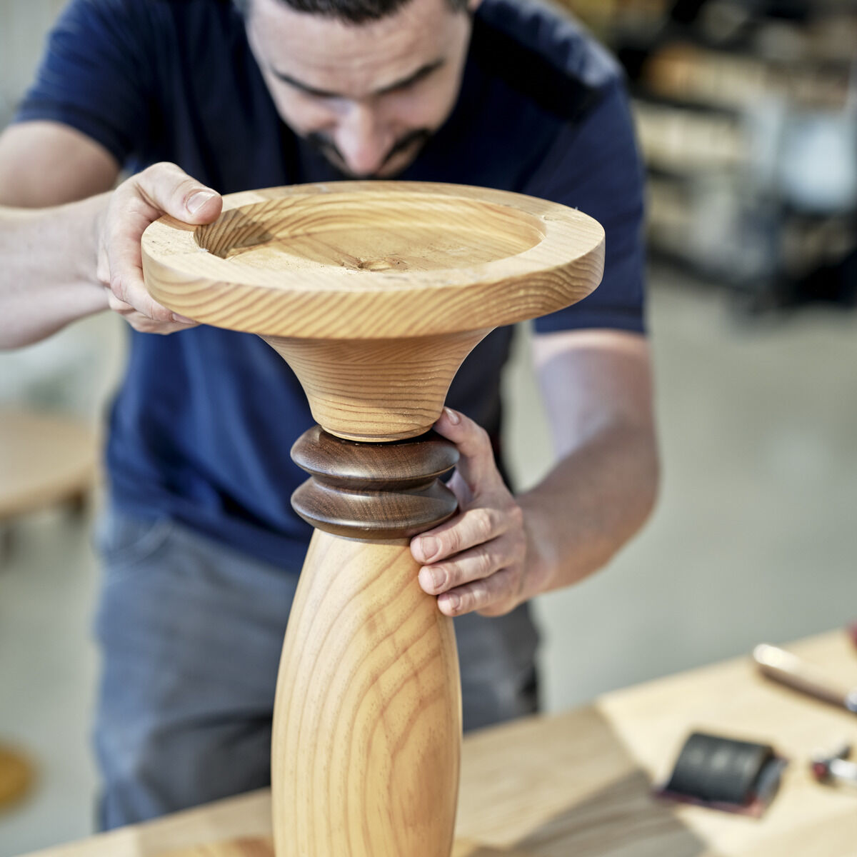 Ein Mann montiert einen klassischen Beistelltisch »Birillo« aus Holz. Der Tisch hat ein elegantes, rustikales Design und besteht aus hellem und dunklem Holz. Auf den Tischbeinen und dem Gestell sind feine handwerkliche Details zu erkennen. Im Hintergrund ist eine Werkstatt zu sehen.
