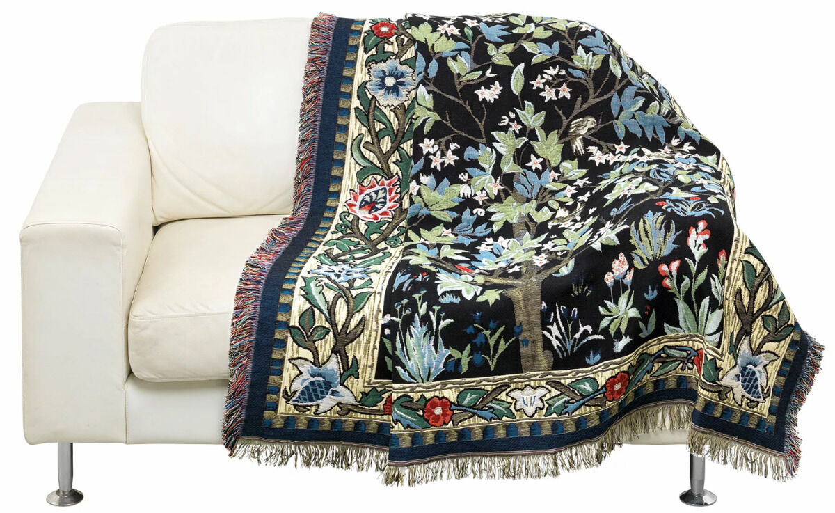 Wohnplaid »Lebensbaum« nach William Morris, drapiert auf einem weißen Sofa. Das Plaid zeigt ein komplexes und farbenfrohes Muster mit Bäumen und Blumen in Schwarz, Grün, Blau, Rot, Creme und Weiß.