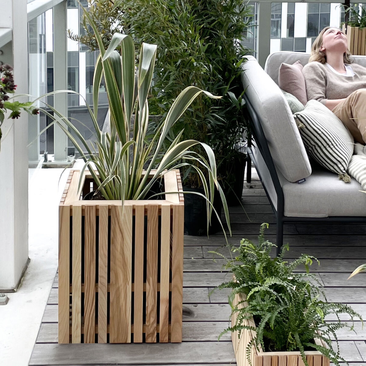 Ein Pflanzgefäß 'GrowBIG' aus Holz steht auf einem Balkon. Es ist mit grünen Pflanzen gefüllt. Im Hintergrund befindet sich eine Sitzecke mit hellen Kissen.