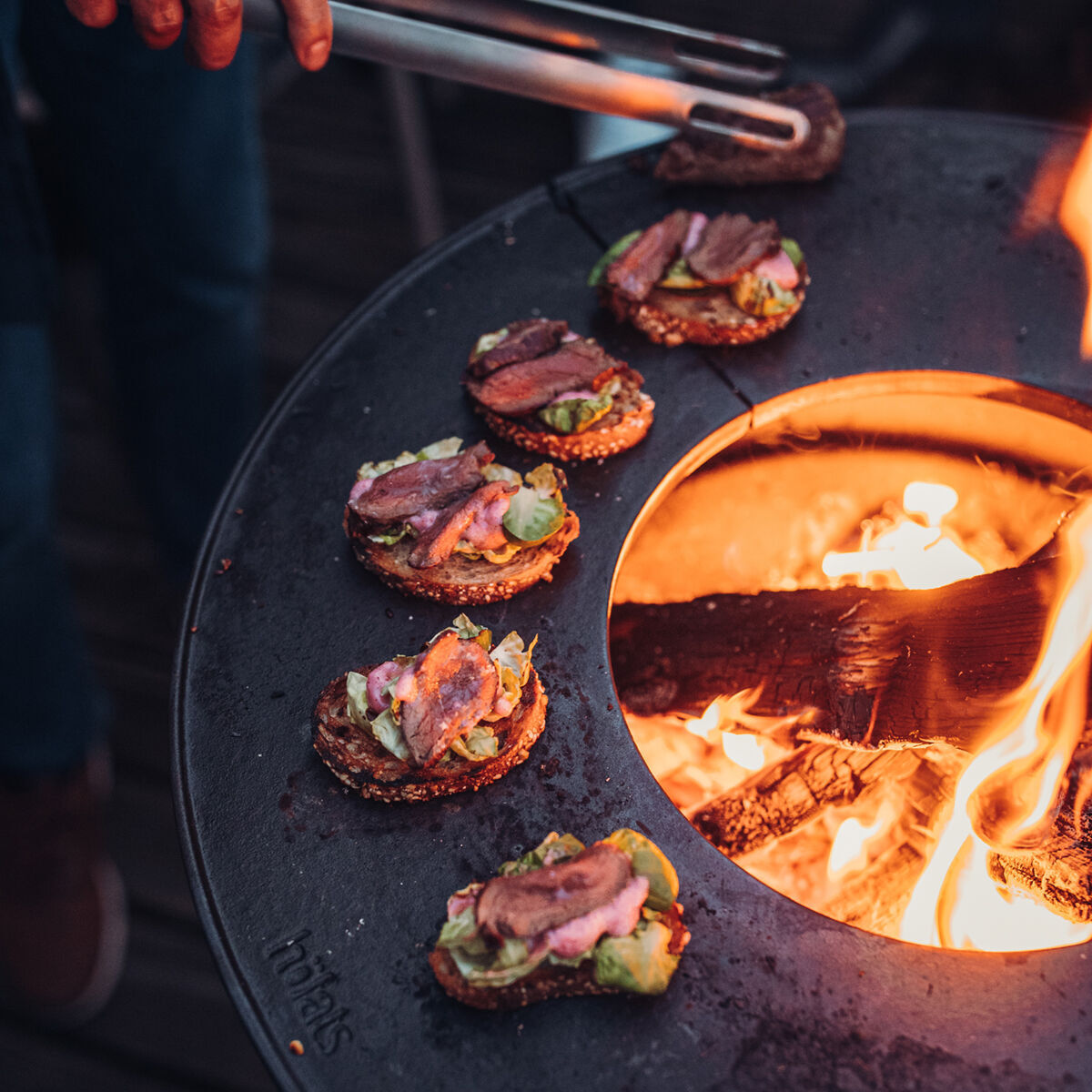 Ein Plancha Halbring Grill, auf dem Fleisch und Gemüse über einem offenen Holzfeuer zubereitet werden. Personen greifen nach Essen auf dem Grill.