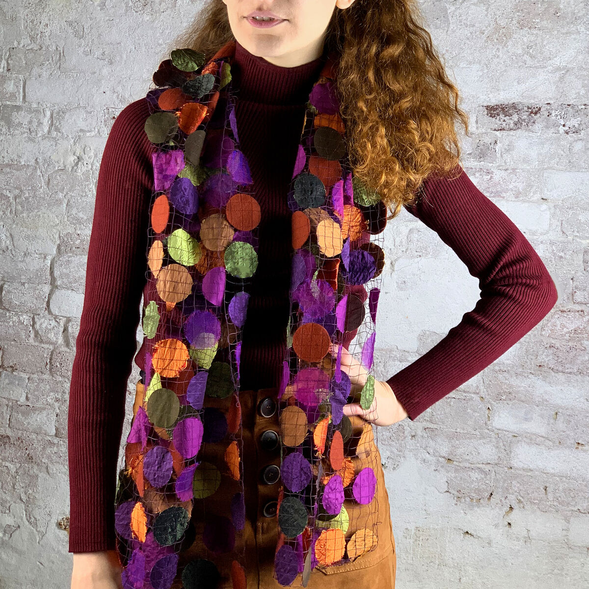 Eine Frau trägt einen transparenten Schal in warmen Gewürztönen, bestehend aus bunten Kreisen in Rot, Orange, Grün, Lila und Braun. Der Schal bietet einen stilvollen Kontrast zu ihrem burgunderroten Pullover und der braunen Hose.