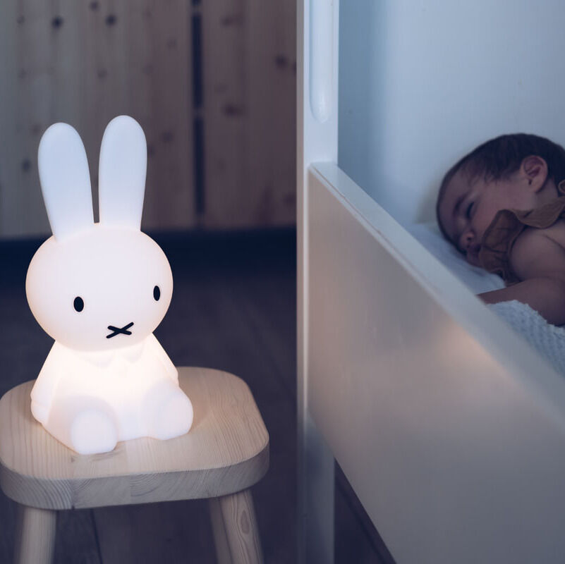 Ein Nachtlicht in Form von Miffy steht auf einem Holzhocker neben einem schlafenden Baby im Kinderbett.