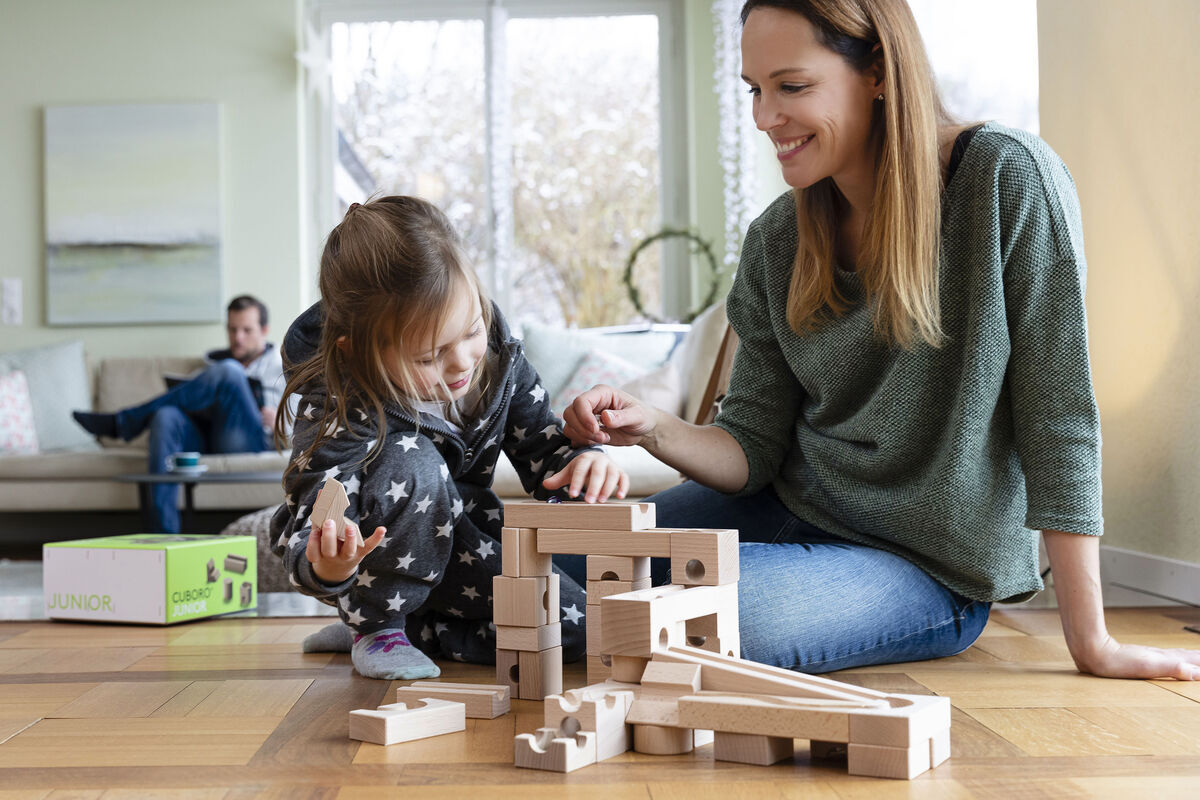 Eine Mutter und ihre Tochter sitzen auf dem Boden und spielen gemeinsam mit einem Holz-Kugelbahn-Bausatz. Im Hintergrund sind weitere Personen auf einem Sofa zu sehen.