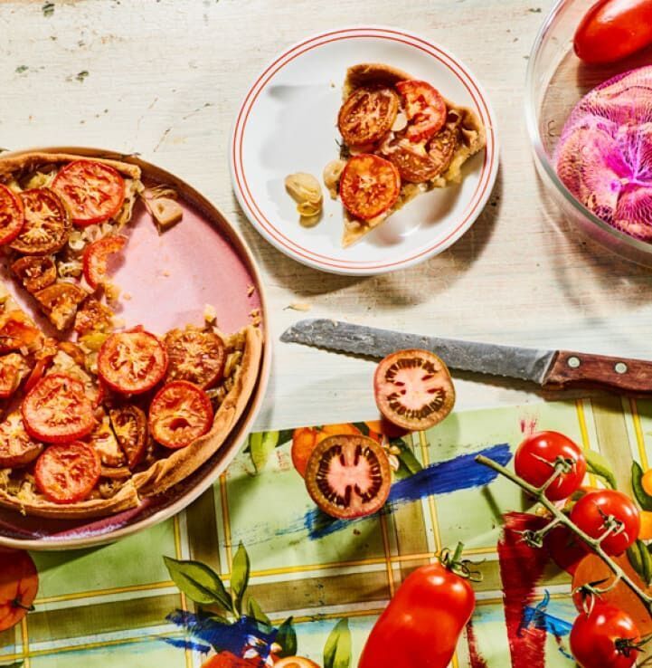 Ein gedeckter Tisch mit Tomatenkuchen auf einem rosa Teller, ein Stück auf einer kleinen weißen Platte, tomatenrote Tomaten an einem Stiel, ein scharfes Messer und eine karierte Tischdecke. Ideal für Wochenmarkt- und Backbuch-Liebhaber.