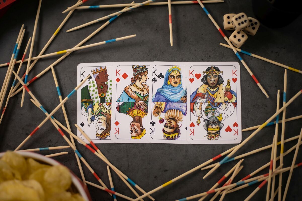 Spielkarten-Set 'Rommé' mit vier Königen, umgeben von Mikado-Stäbchen und Würfeln auf einem Tisch.