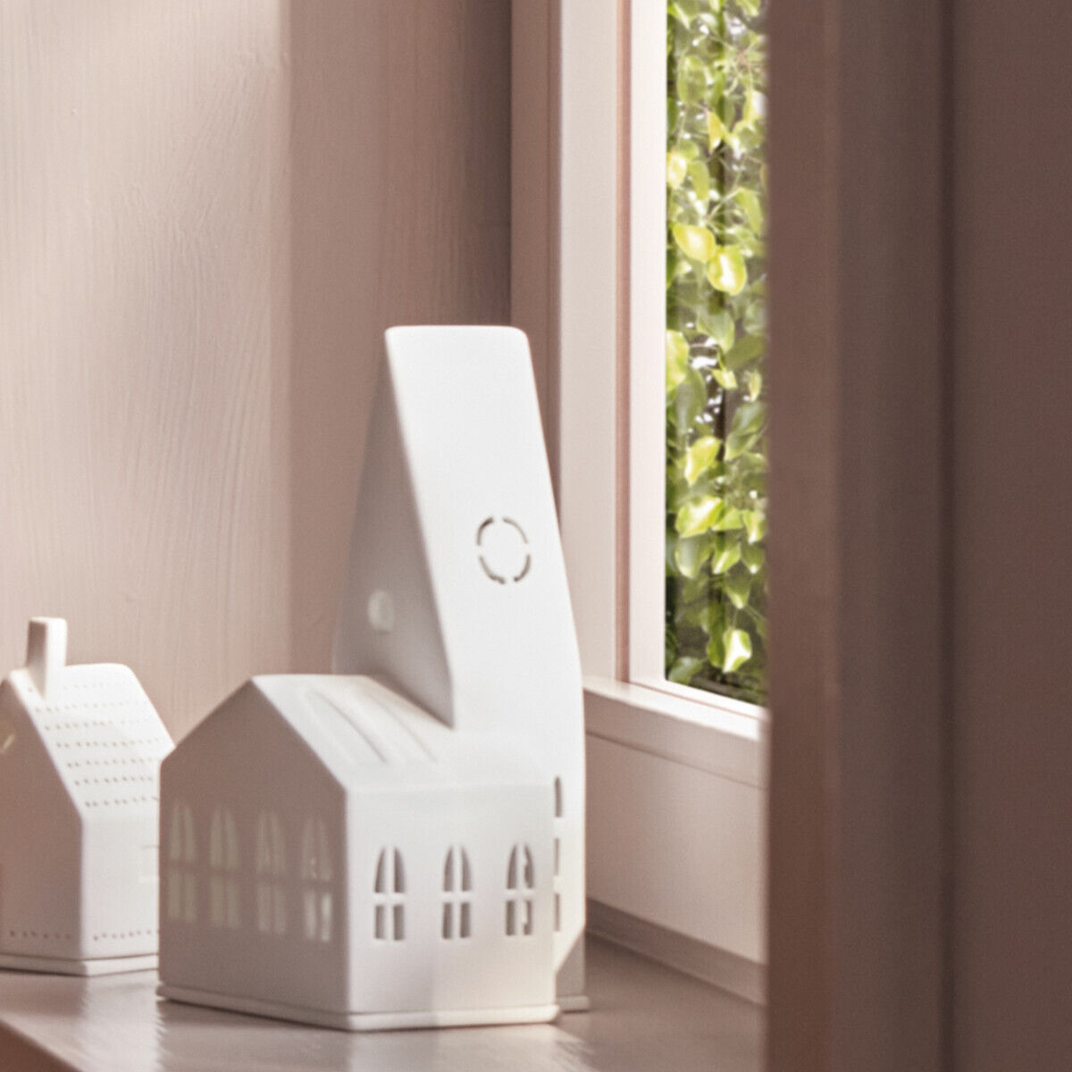 Weiße Keramiklichthäuser in Form von kleinen Kirchen, die auf einer Fenstersims stehen. Sie strahlen ein warmes und gemütliches Licht aus, mit einem Hintergrund aus grünen Pflanzen, die durch das Fenster zu sehen sind.