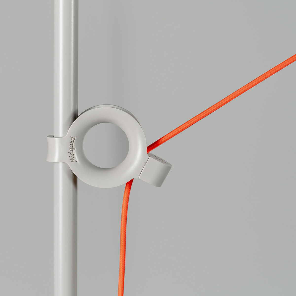 Nahaufnahme einer Designer-Stehlampe »AYNO« mit einem weißen Rahmen und einem orangefarbenen Kabel, das um einen runden Halter gewickelt ist, vor einem grauen Hintergrund.