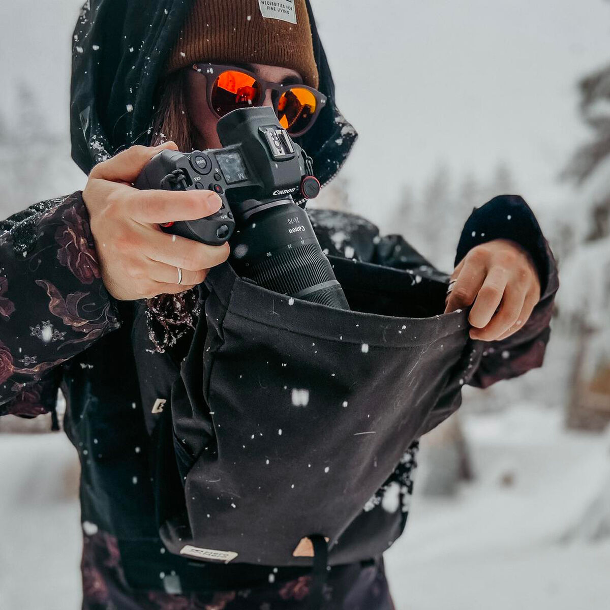 Eine Person in Winterkleidung mit einer braunen Mütze und einer schwarzen Jacke steht in einer verschneiten Landschaft und hält eine schwarze Tasche »Piha«, in der sich eine Kamera befindet. Es schneit leicht.