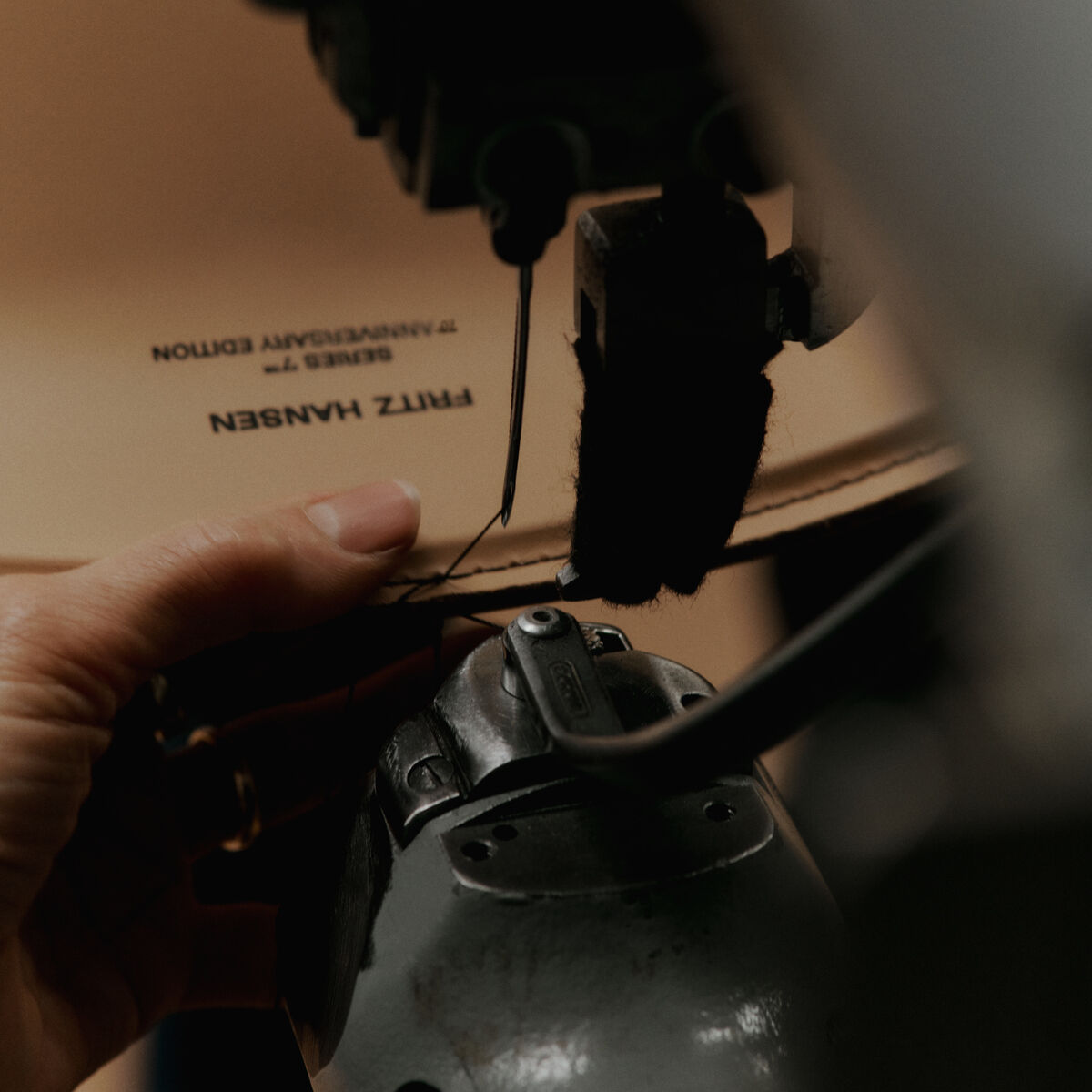 Detailaufnahme einer Hand, die sorgfältig einen Lederbezug an einer Nähmaschine verarbeitet. Auf dem Leder ist die Aufschrift 'FRITZ HANSEN SERIES 7™ ANNIVERSARY EDITION' zu erkennen. Die Szene zeigt den präzisen Herstellungsprozess eines Designerstuhls in warmen, natürlichen Farbtönen.