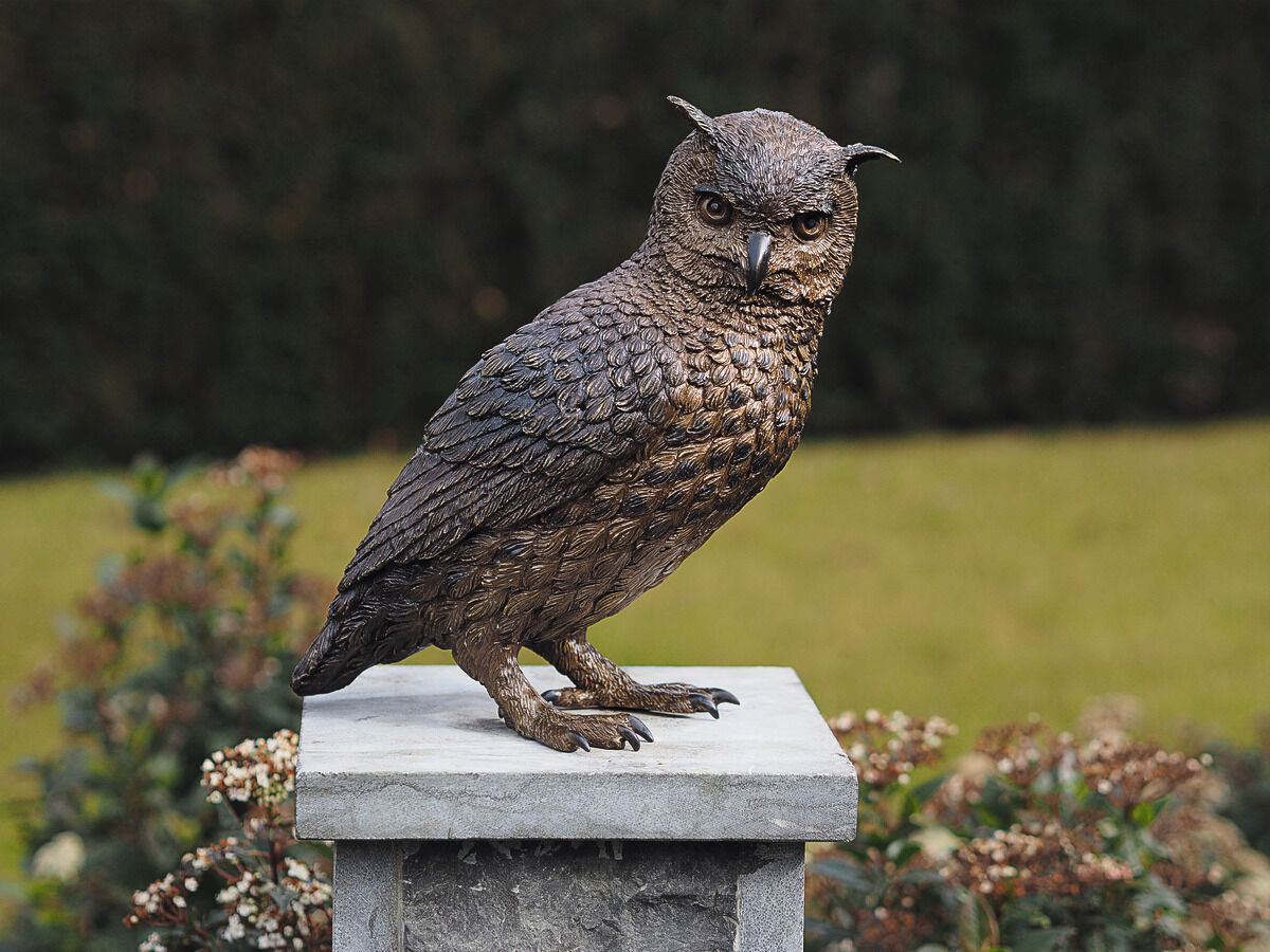 Eine naturgetreue Gartenskulptur in Form einer bronzenen Eule sitzt auf einem grauen Steinsockel. Im Hintergrund ist unscharfes Grün von Pflanzen und Rasen zu sehen.