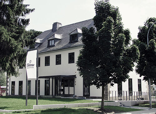 Modernes weißes Bürogebäude mit schwarzem Dach, umgeben von Bäumen und einem Schild mit dem Schriftzug 'Tutima'