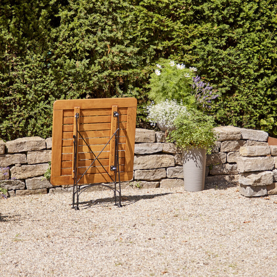 Ein zusammengeklappter Holztisch steht auf einem Kiesboden vor einer Steinmauer und grünem Gebüsch. Neben dem Tisch befindet sich ein großer grauer Blumentopf mit weißen und lila Blumen.