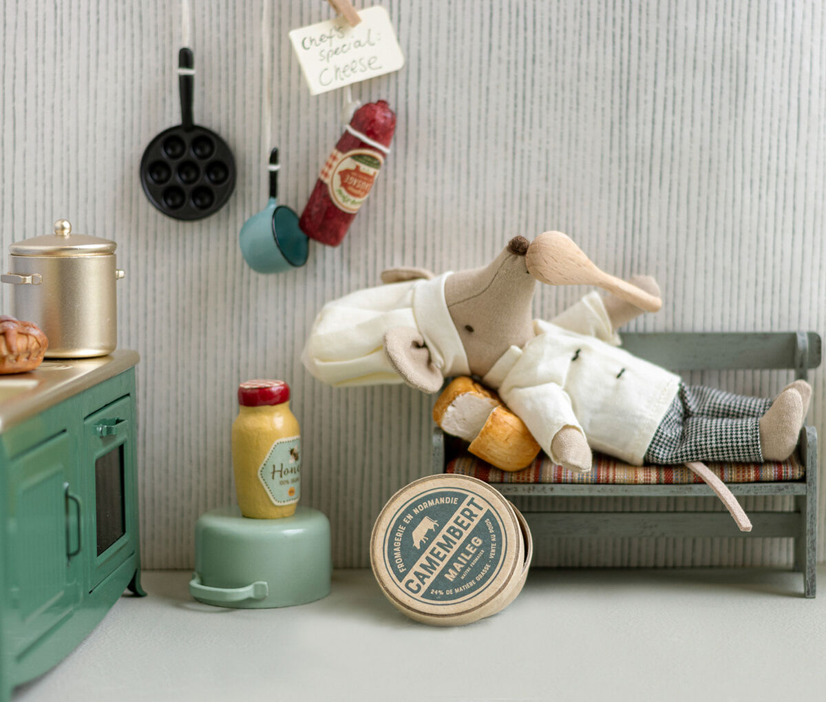 Eine niedliche Stoffmaus in Kochkleidung liegt auf einer kleinen Bank neben einer Miniaturküche mit Suppentopf und Löffel. Im Hintergrund sind Küchenutensilien, Camembert- und Senfdose sowie Wurstdeko zu sehen. Die Szene vermittelt ein charmantes Küchen- und Kochthema, ideal als Dekoration oder Spielzeug.