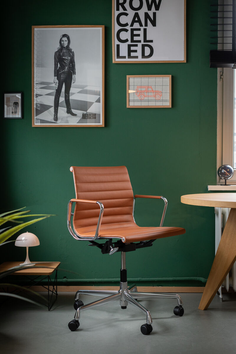 Vitra Alu Chair EA 118, ein polierter, ergonomischer Bürostuhl aus braunem Leder und Aluminiumrahmen, steht vor einer grünen Wand mit gerahmten Bildern. Ein modernes Büro-Setup mit einem Schreibtisch, einer Lampe und Pflanzen ist sichtbar.