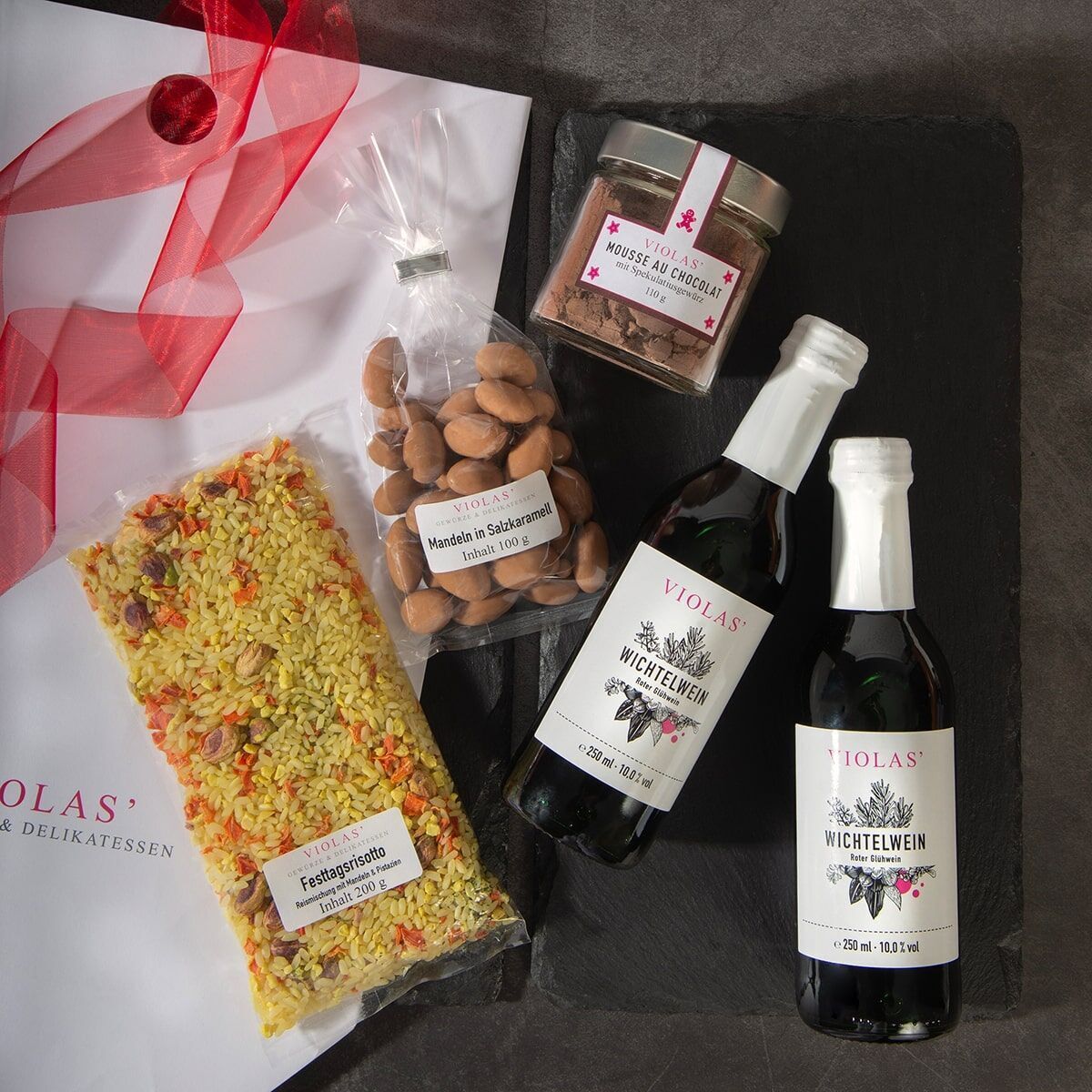 Ein Weihnachtszeit-Set bestehend aus zwei Flaschen Wichtelwein, einer Packung festlichen Reis, Schokoladenmandeln und einem Glas Schokolade-gebrochenem Mandelstücken, präsentiert in einer Geschenkverpackung mit rotem Band.