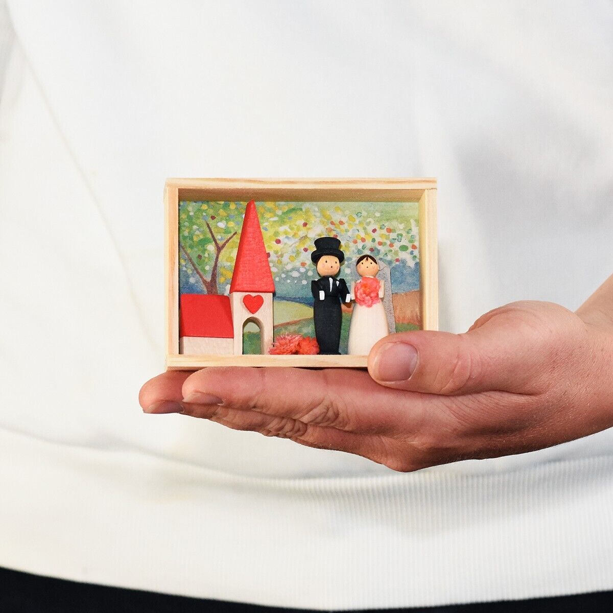Eine hölzerne Miniaturszene, die ein Brautpaar in einer kleinen, handgehaltenen Holzbox darstellt. Im Hintergrund sind eine rote Kirche und ein Herz zu sehen, sowie Bäume und ein blauer Himmel.