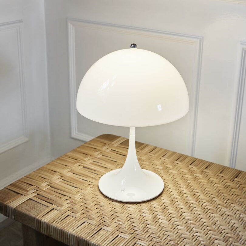 Die Leuchte »Panthella 250 Portable« aus Acryl in Opal Weiß steht auf einem Tisch mit geflochtener Oberfläche. Die moderne Designlampe strahlt Weichheit und Eleganz aus und passt ideal in verschiedene Wohnbereiche wie Wohnzimmer oder Schlafzimmer.