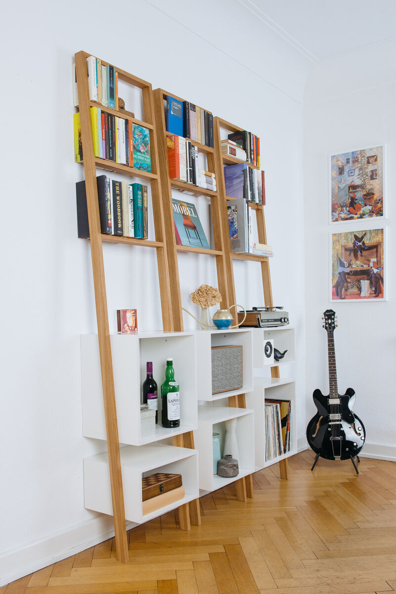 Ein modernes Leiterregal namens »brecht« in einem Wohnzimmer. Das Regal besteht aus einem weißen Regalteil und einem natürlichen Holzgestell. Es ist mit verschiedenen Büchern, Dekorationsgegenständen und einer Gitarre dekoriert. Das Regal steht auf einem Parkettboden, und an der weißen Wand hängen Bilderrahmen.