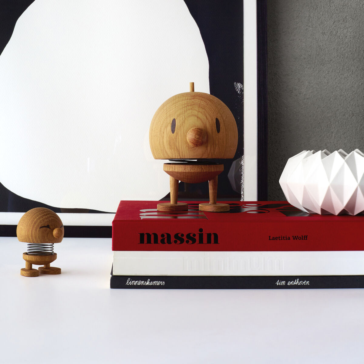 Ein Hoptimist Bumble Eiche in Größe S steht auf einem Buchdeckel neben einer kleineren Hoptimist-Figur. Im Hintergrund ist ein minimalistisches, schwarz-weißes Kunstwerk zu sehen.