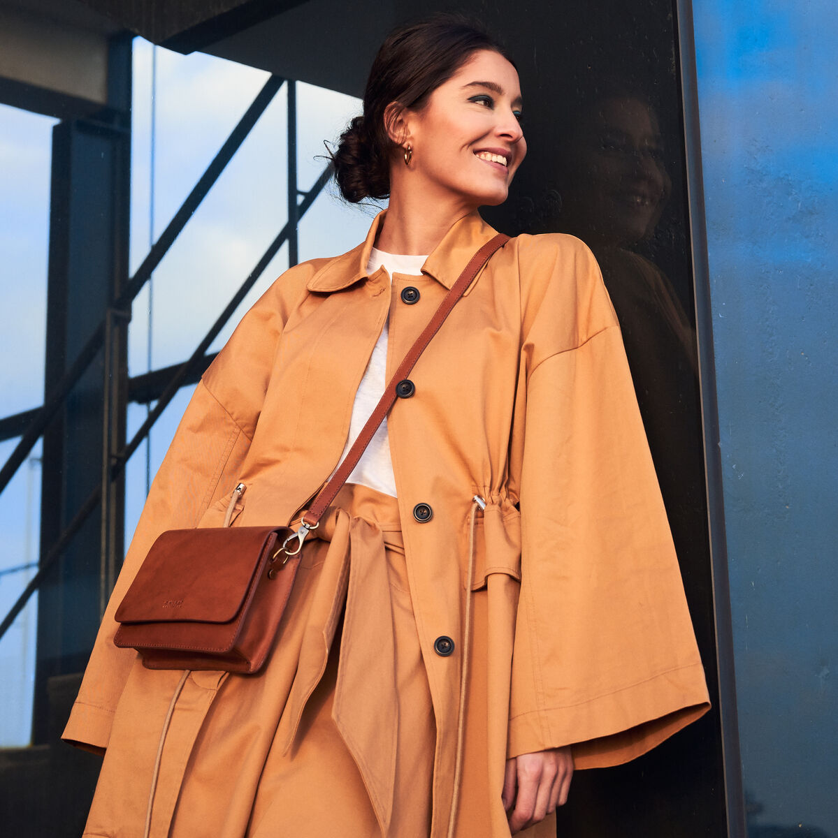 Frau trägt eine braune Apfelledertasche namens 'Audrey Mini', kombiniert mit einem stylischen Trenchcoat, vor einer modernen Architekturkulisse.