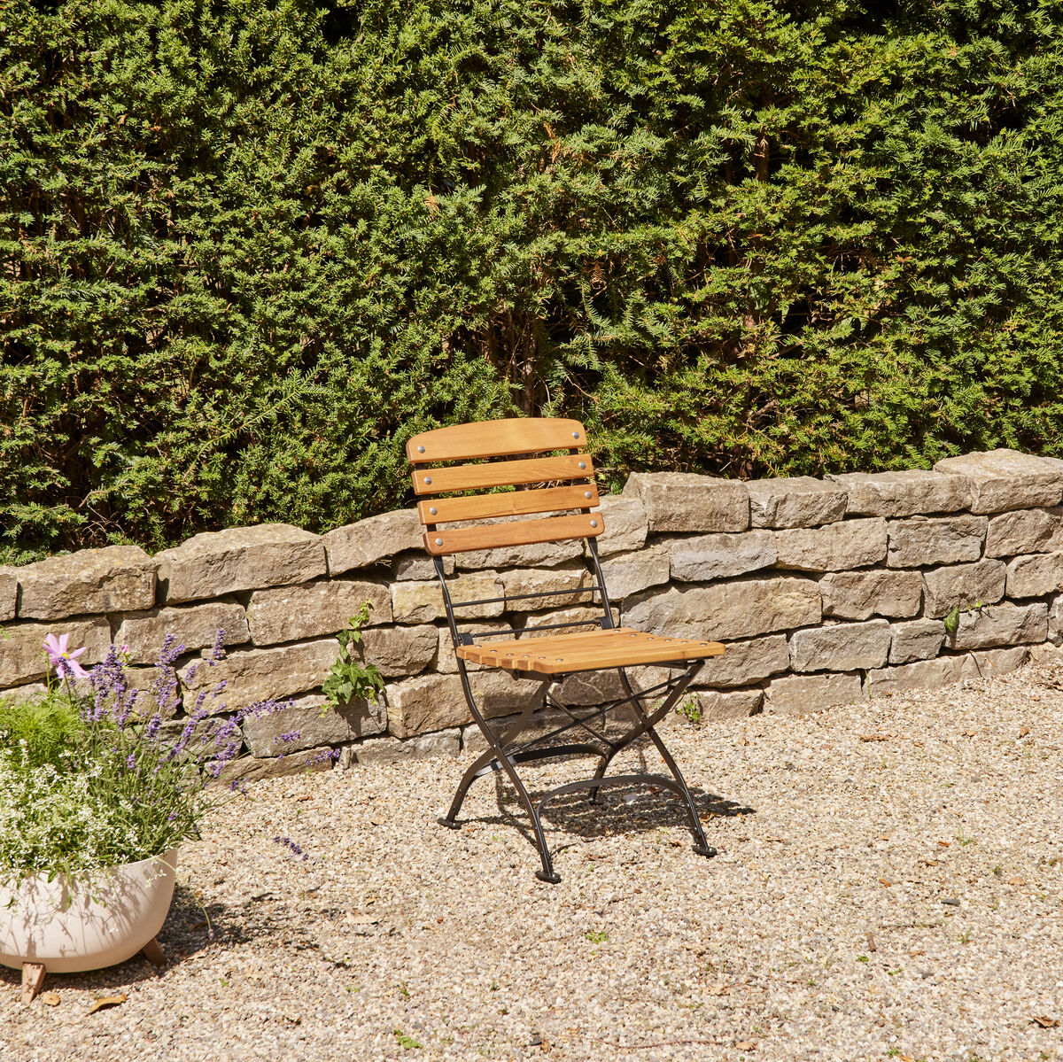 Ein Klappsessel aus Holz und Eisen steht auf einer Kiesfläche im Garten, umgeben von Pflanzen und grünem Heckengebüsch.