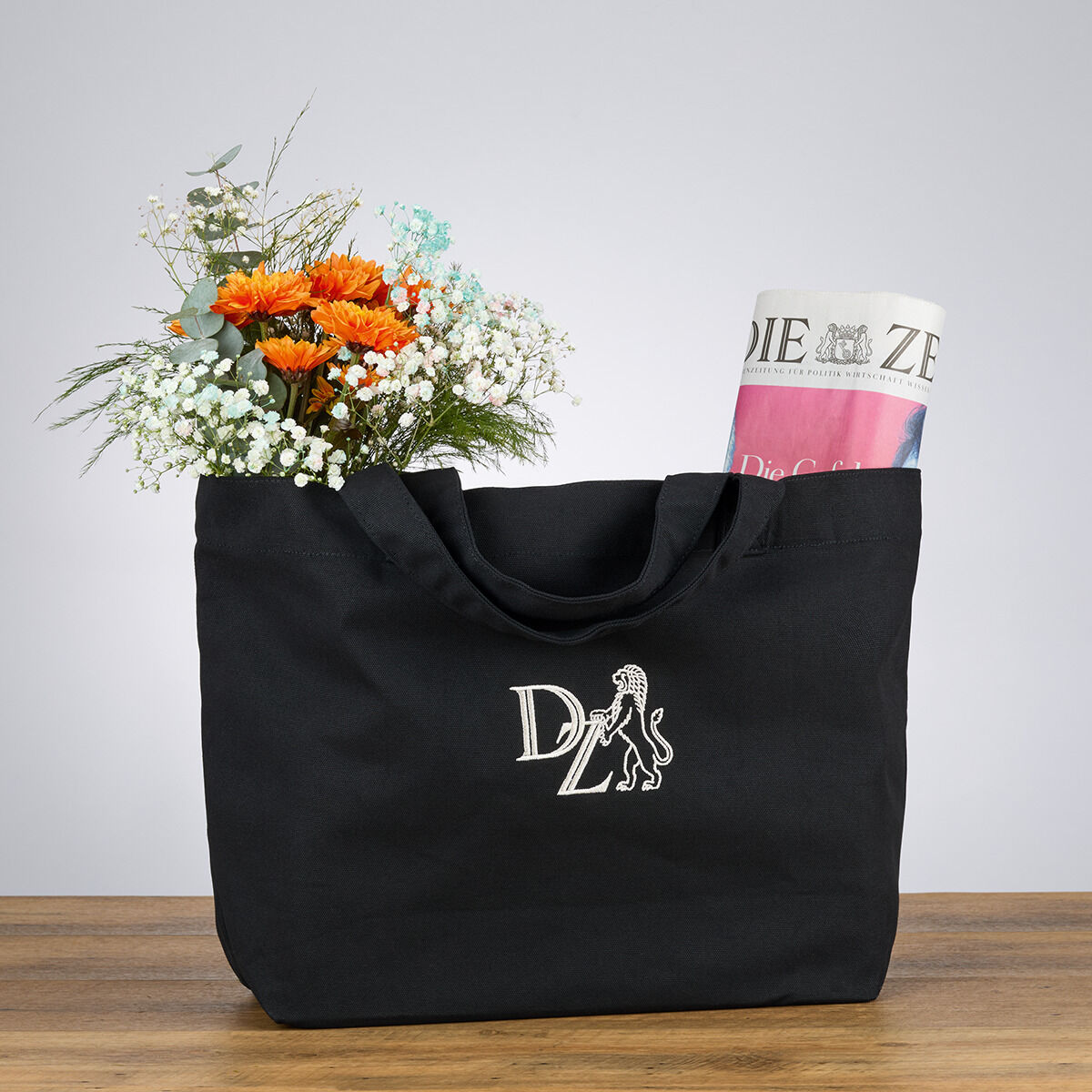 Schwarze Canvas-Tasche mit ZEIT-Logo auf einem Holztisch, gefüllt mit einer Zeitung und einem Strauß Blumen.