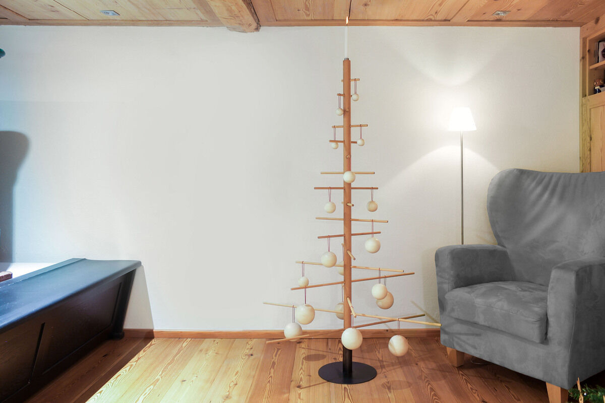 Ein abstrakter Weihnachtsbaum aus Holz mit einem Stahlständer steht in einem modern eingerichteten Wohnzimmer. Der Baum ist mit minimalistischen Kugelornamenten geschmückt und steht neben einem grauen Sessel. Die Wände sind weiß gestrichen, und der Boden besteht aus Holzplanken.