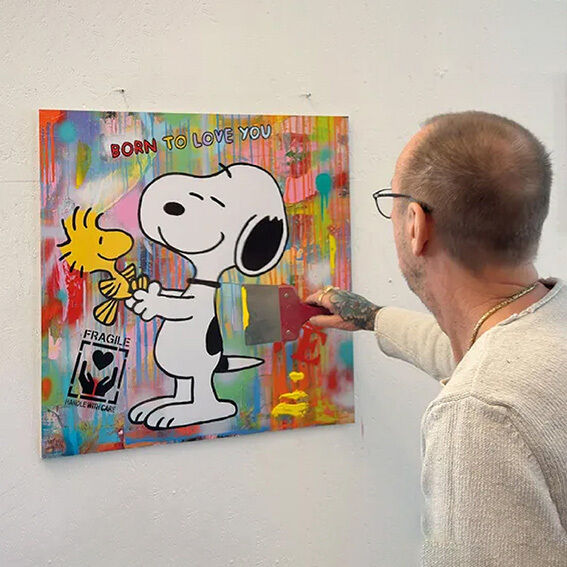 Das Bild zeigt einen Künstler, der an einem farbenfrohen Pop-Art-Kunstwerk arbeitet. Es zeigt Snoopy und Woodstock mit dem Spruch "BORN TO LOVE YOU". Der Hintergrund ist bunt mit Sprühfarbe gestaltet. Das Werk hat einen modernen, individuellen Charakter und ist 70 x 70 cm groß.