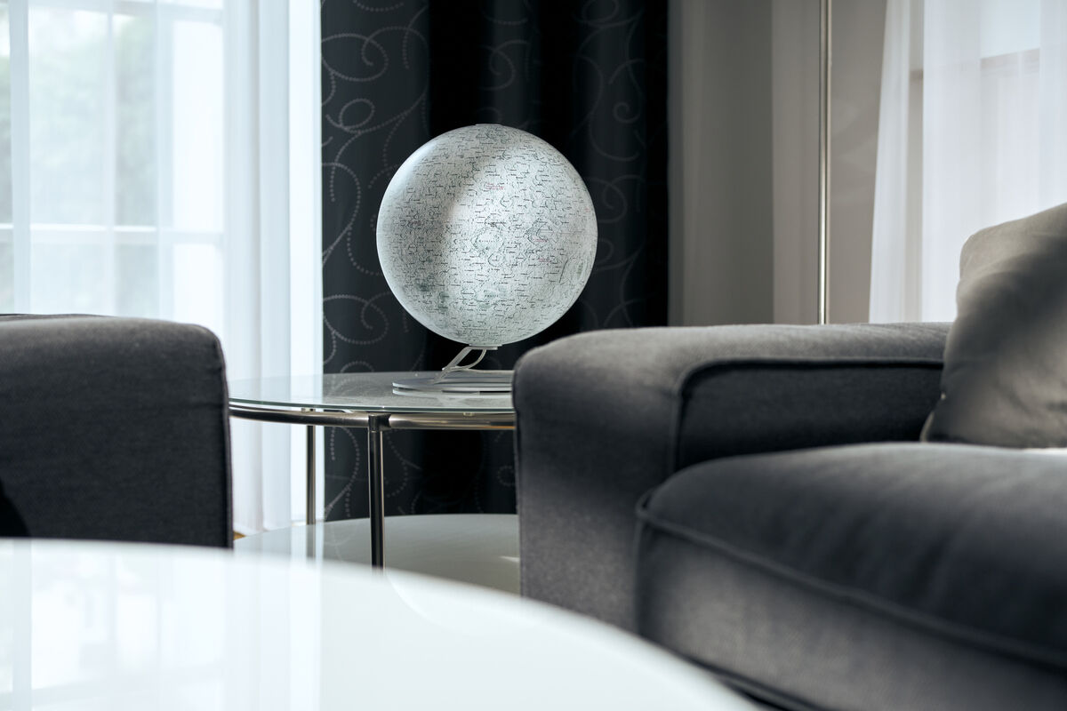 Ein Leuchtglobus von National Geographic mit der Darstellung des Mondes steht auf einem runden Glastisch zwischen zwei grauen Sofas vor einem Fenster.