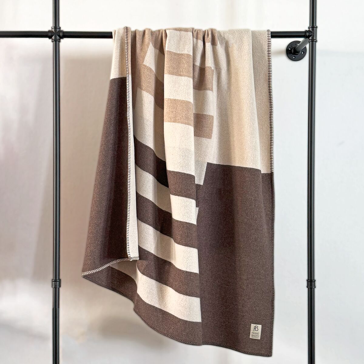 Eine hochwertige Kaschmirdecke der Marke Eagle Products hängt locker über einer schwarzen Metallstange. Die Decke zeigt ein dezentes, modernes Blockstreifenmuster in Naturtönen von Creme, Naturweiß bis Dunkelbraun und Hellbraun. Die Materialstruktur erscheint weich und sehr fein, die Ränder sind sorgfältig eingefasst.