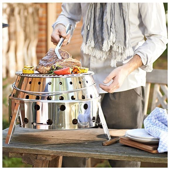 Eine Person grillt Fleisch und Gemüse auf einem silbernen Grill »Atago« aus rostfreiem Stahl im Freien. Der Grill steht auf einem Tisch aus Holz neben einem Geschirrtuch und Teller.