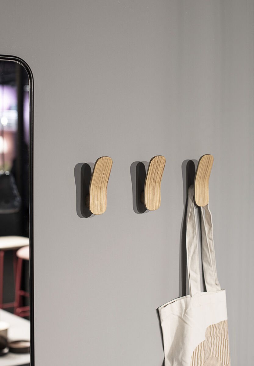 Drei Kleiderhaken aus Eiche, montiert an einer hellgrauen Wand. An einem Haken hängt eine helle Stofftasche. Die Haken haben ein minimalistisches und modernes Design.