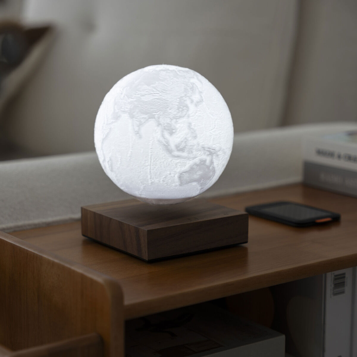Die Gingko Design »Smart Earth Lamp« schwebt leuchtend über einem Holzboden auf einem elegantem Beistelltisch im Wohnzimmer. Im Hintergrund befindet sich ein Sofa, Bücher und ein Smartphone ergänzen das stilvolle, moderne Ambiente.