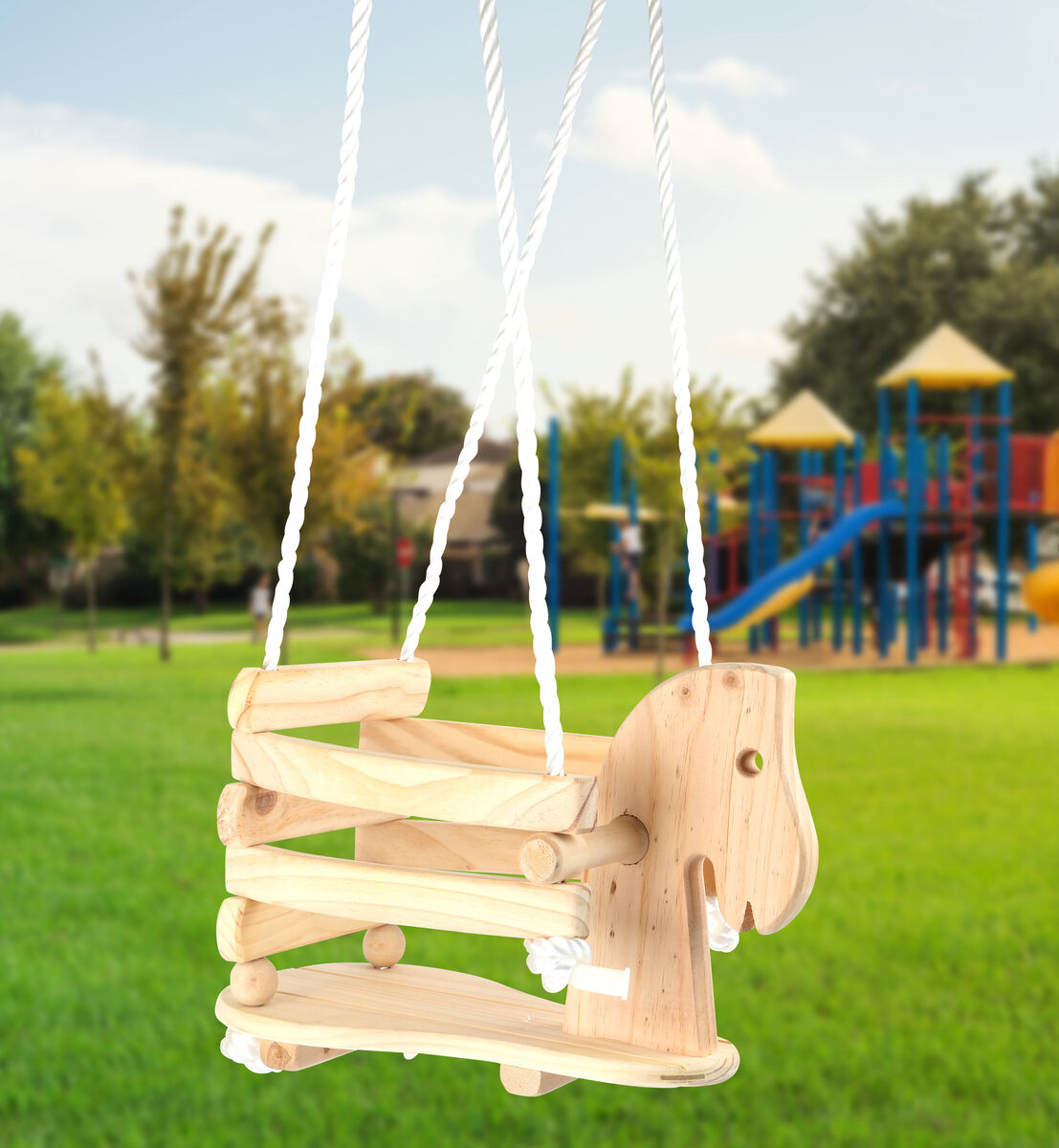 Holzschaukel in Form eines Ponys hängt an weißen Seilen im Freien. Im Hintergrund befindet sich ein Spielplatz mit Spielgeräten und grünem Rasen.
