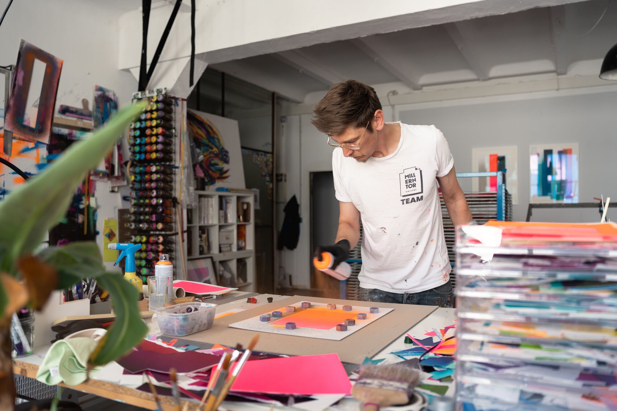 Junger Mann in weißem T-Shirt arbeitet an einem Kunstprojekt in einem Atelier mit vielen Farben und Papieren