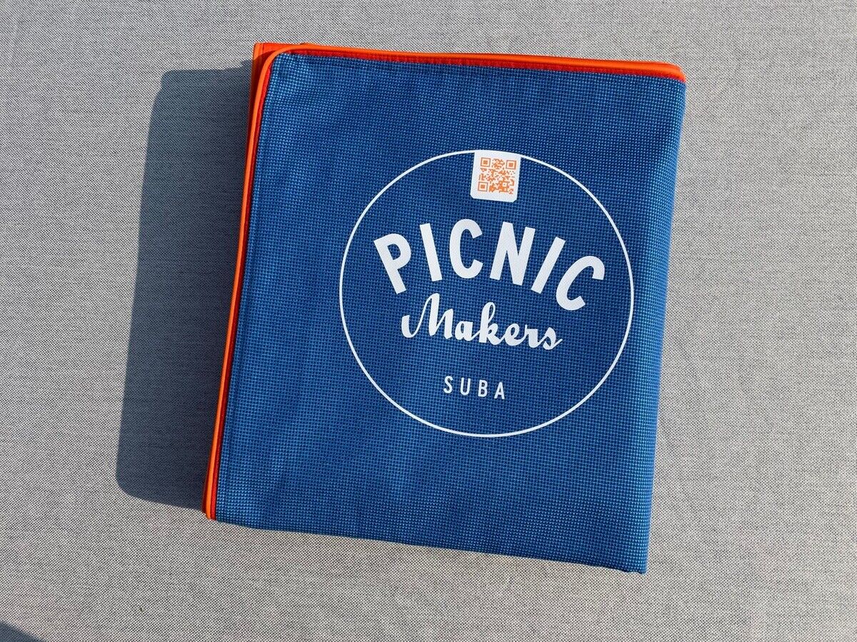 Eine zusammengefaltete blaue Picknickdecke mit dem Aufdruck 'Picnic Makers Suba' und einem Rundlogo in weiß. Die Kanten der Decke sind orange umrandet und sie liegt auf einer grauen Oberfläche.