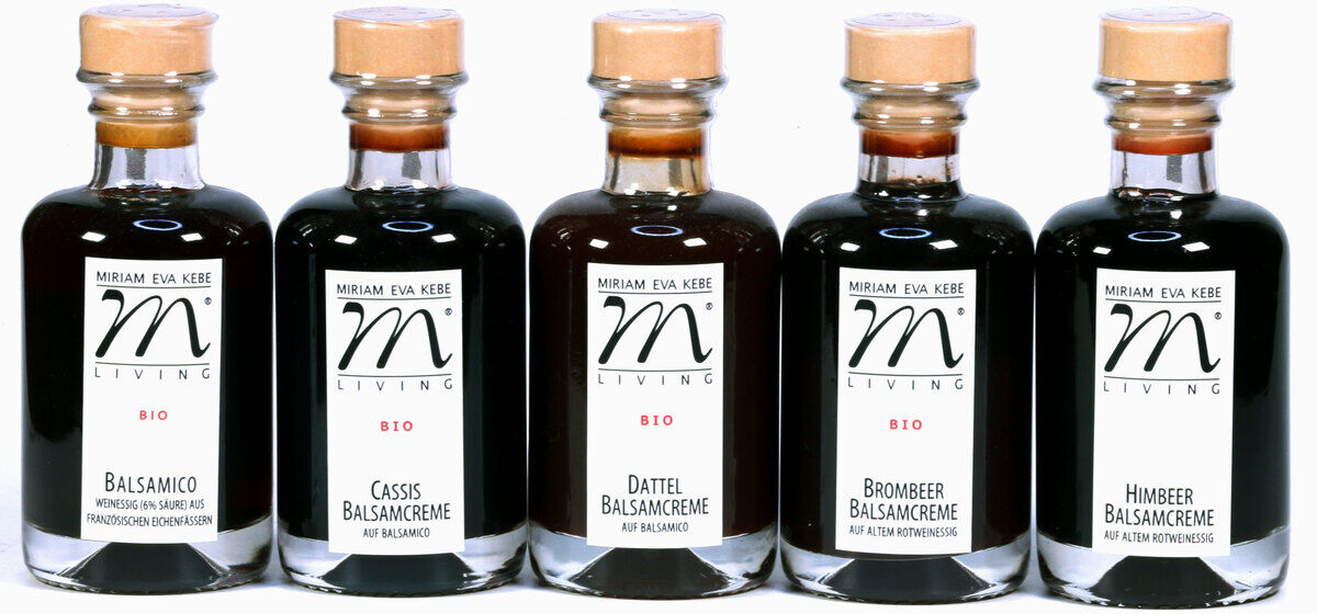 Das Bild zeigt ein Set von fünf Flaschen mit verschiedenen Balsamico-Essigen. Die Flaschen haben dunkle Flüssigkeiten und sind mit braunen Korken verschlossen. Auf den Etiketten sind die Namen der Essigsorten und das Logo der Marke zu sehen.