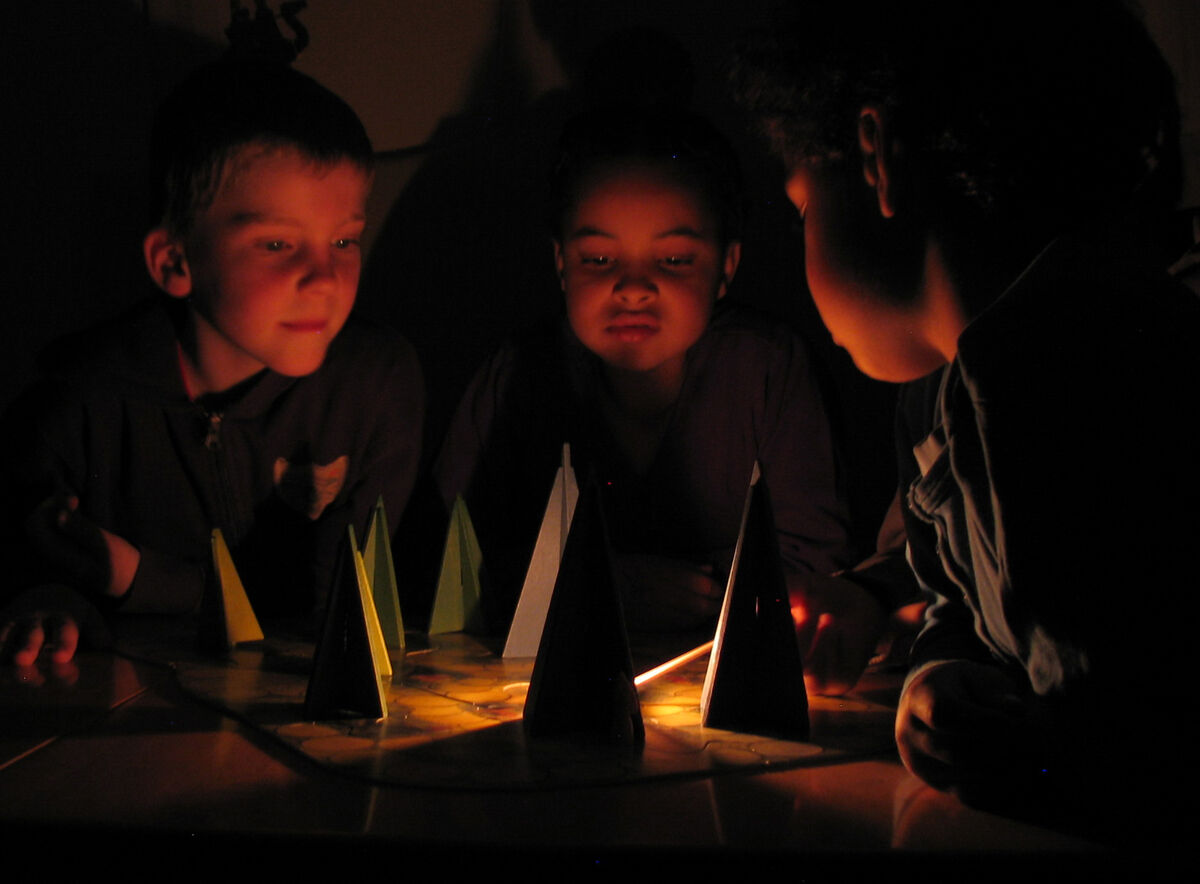 Drei Kinder spielen im Dunkeln das Brettspiel 'Waldschattenspiel', bei dem bunte Pyramidenformen durch eine Kerze beleuchtet werden.