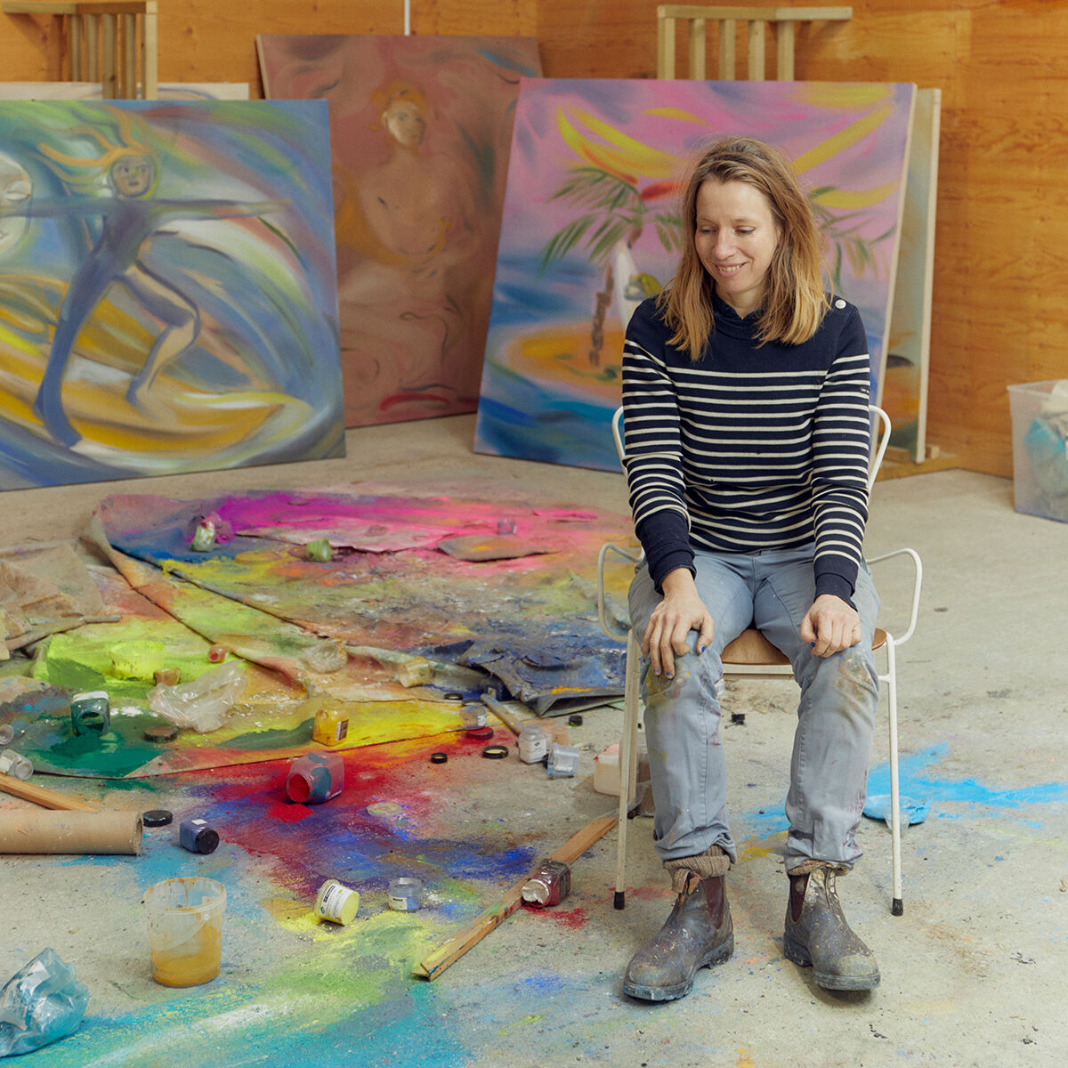 Eine Frau sitzt in einem Atelier vor einer farbigen Leinwand mit abstrakten Kunstwerken. Der Boden ist mit Farben und Malutensilien bedeckt. Die Frau trägt eine gestreifte Oberbekleidung und Jeans.