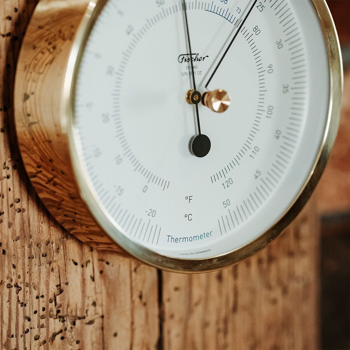 Analoges Thermometer mit goldfarbenem Rahmen und weißem Zifferblatt, montiert an einer Holzoberfläche. Die Skalierung zeigt Temperaturwerte in Grad Celsius.