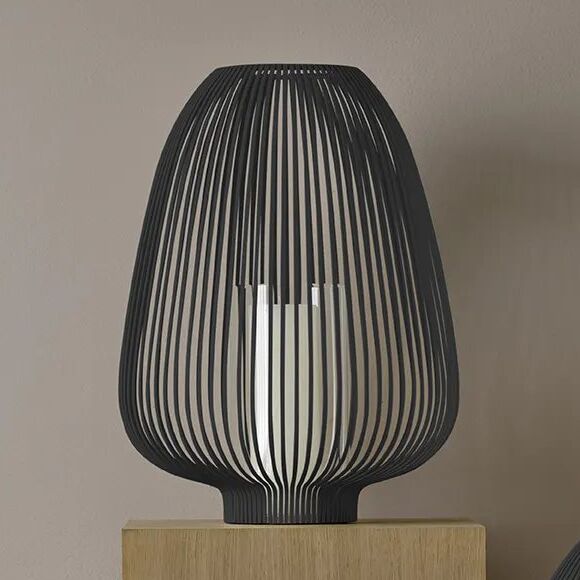 Moderne, schwarze Laterne aus Metall mit schlanken, vertikalen Streben steht auf einem hellen Holzquader vor einer beigen Wand. Die offene Struktur der Laterne zeigt ein zylindrisches Element im Inneren und eignet sich ideal als Wohnungseinrichtung oder stimmungsvolle Dekorationsbeleuchtung.