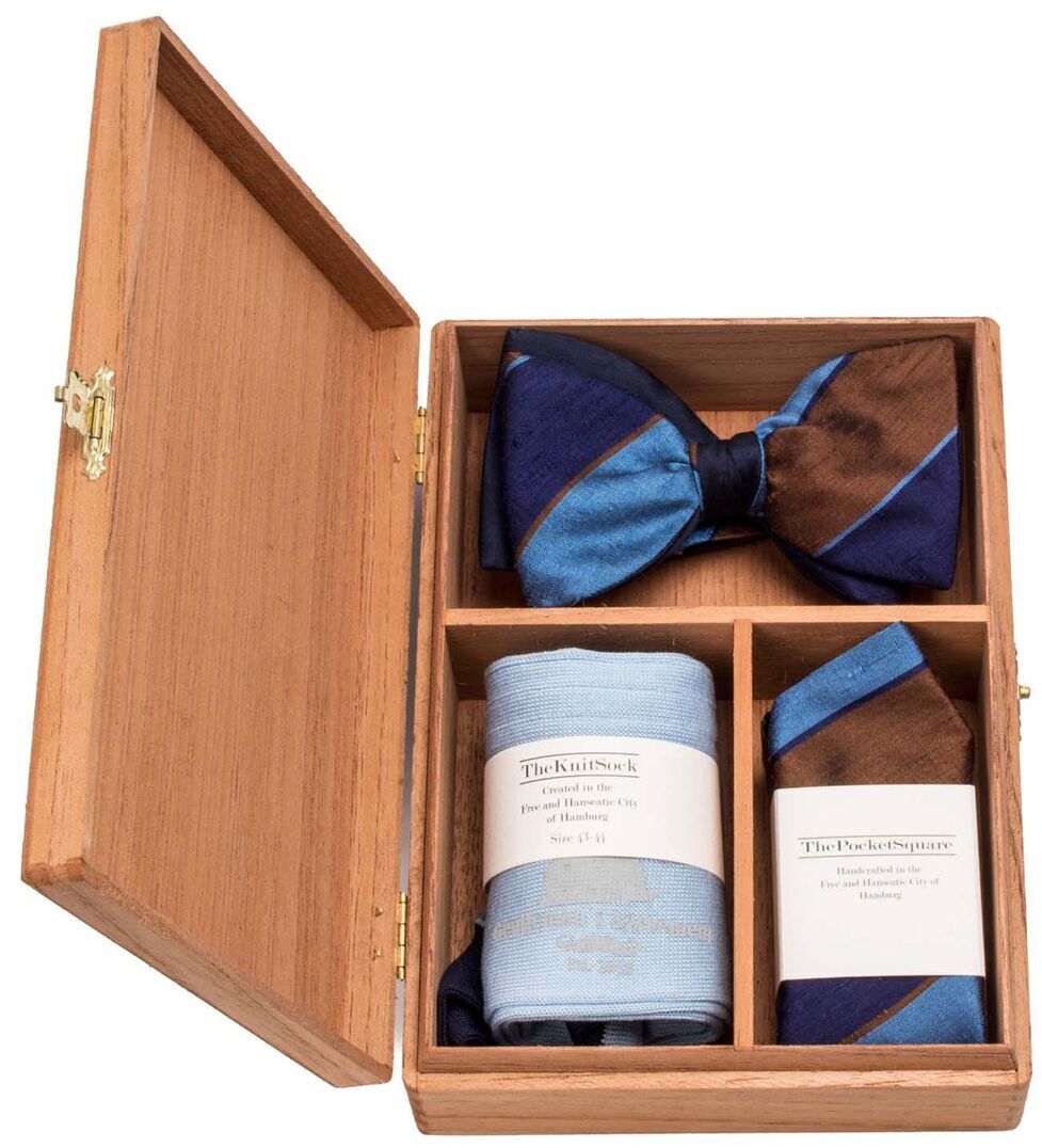 Ein Accessoire-Set namens 'Goldenstripe's Edition' in einer Holzkiste. Enthalten sind eine blaue Fliege mit braunen Streifen, eine blaue Krawatte und ein blau-braunes Einstecktuch. Das Set ist elegant und für formelle Anlässe geeignet.