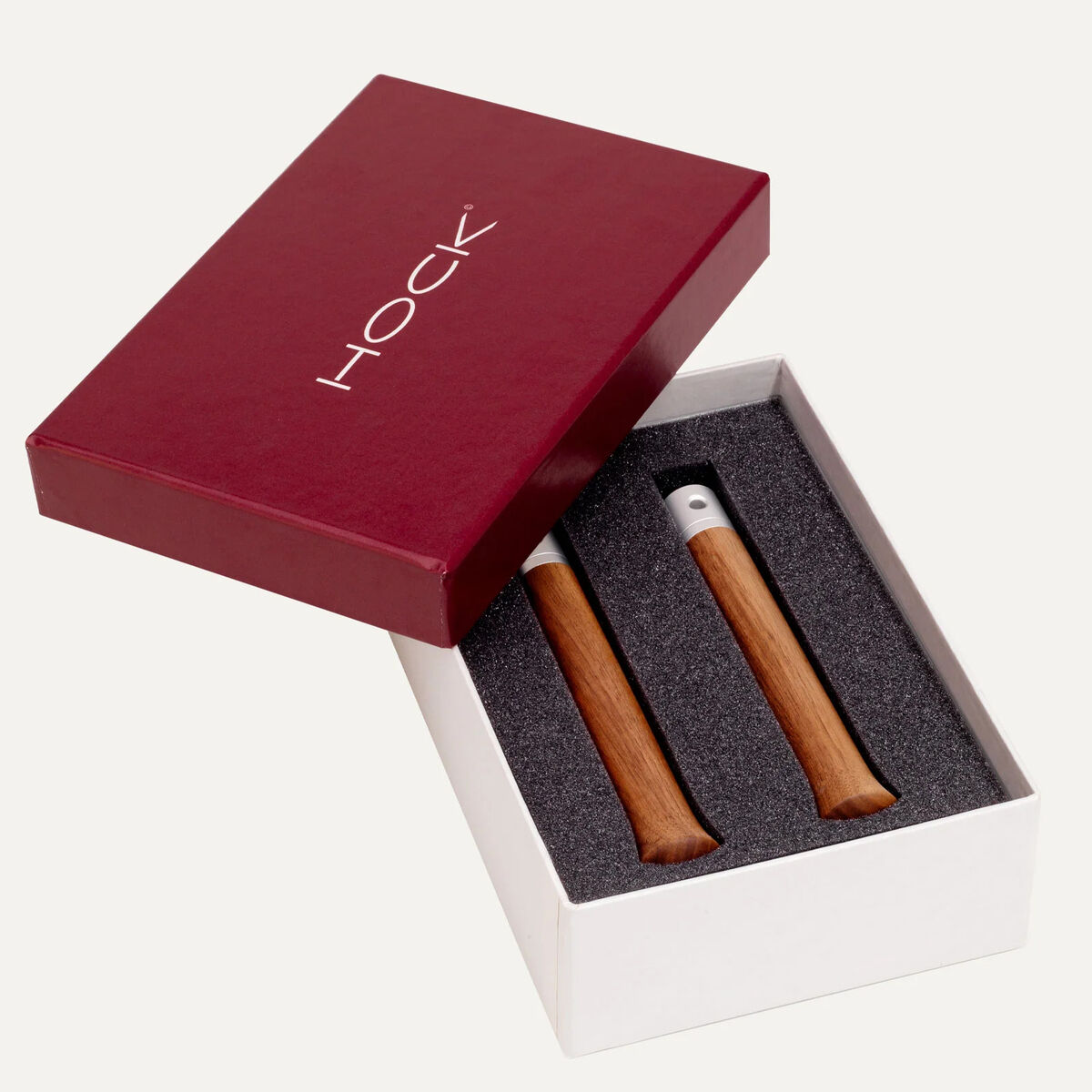 Das Bild zeigt eine elegante Geschenkbox mit dem HOCK-Logo. Im Inneren, eingebettet in schwarzem Schaumstoff, befinden sich zwei fein gearbeitete Holzgriffe aus Nussbaum und Aluminium des Springseils »ROTATOR« von HOCK. Die Box wirkt hochwertig und stilvoll.