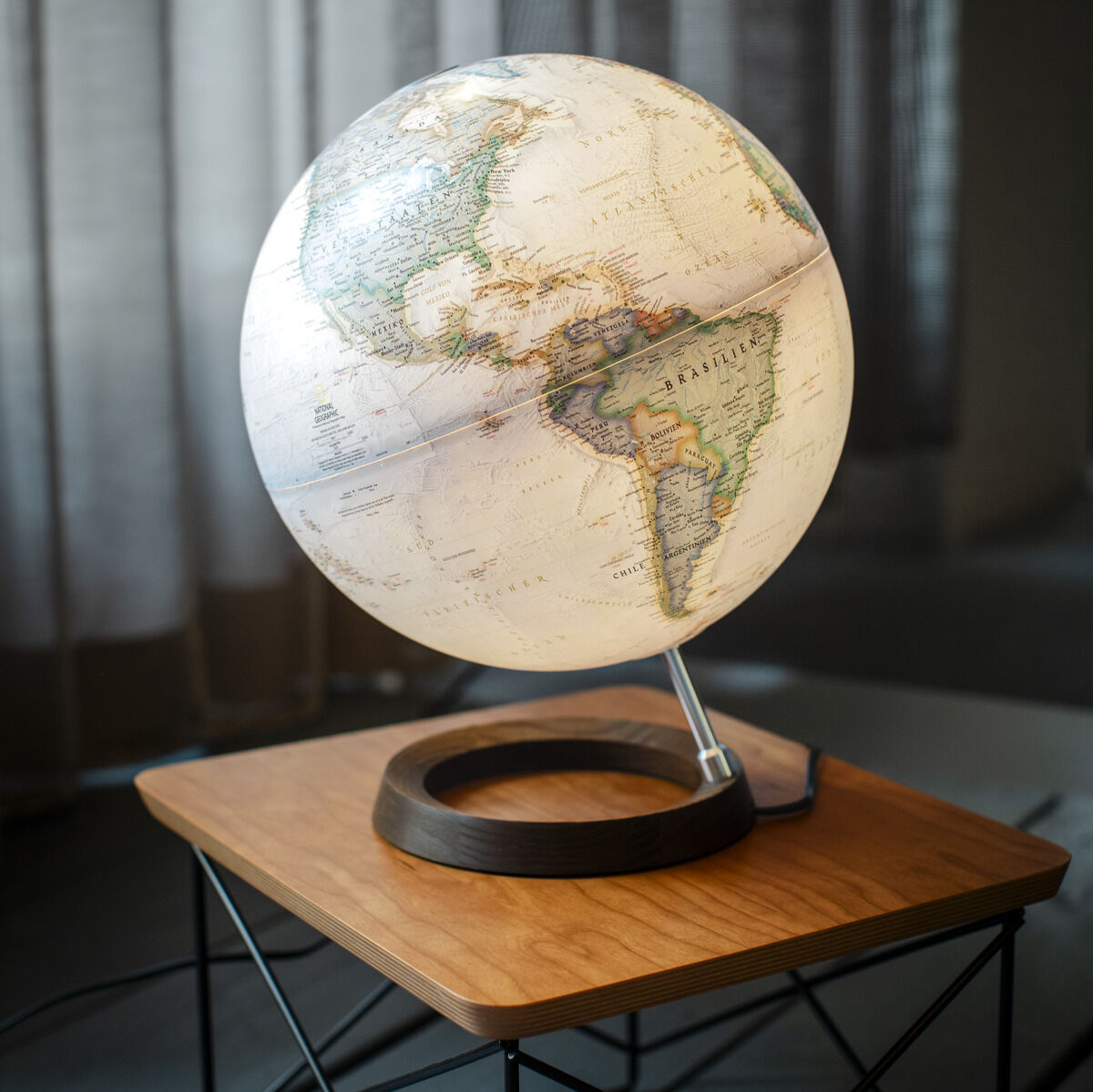 Globus der ZEIT-Sonderedition von National Geographic, beleuchtet, mit detaillierter Weltkarte in neutralen Farben, platziert auf einem modernen Holzständer mit schwarzem Metallgestell, auf einem Tisch vor einem Hintergrund aus Vorhängen.