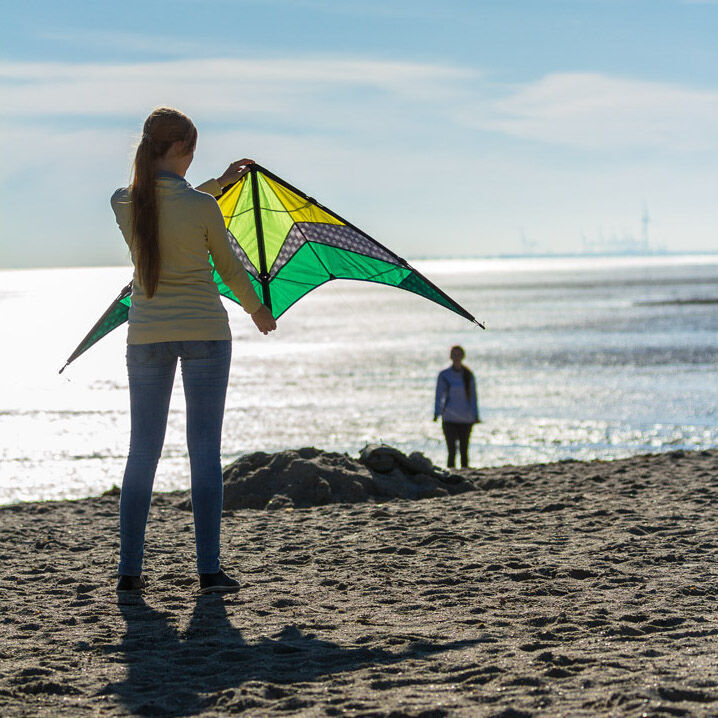 Eine Frau hält einen grünen und gelben Lenkdrachen namens "Limbo II Emerald" am Strand, während eine weitere Person im Hintergrund entlang des Meeres steht und der Himmel klar ist.