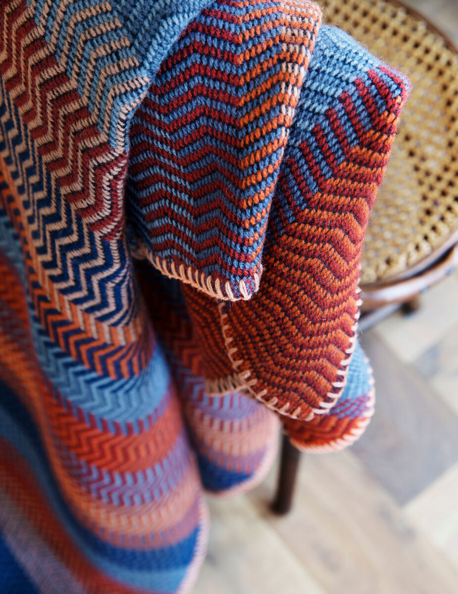 Bunte Lammwolldecke mit Zigzag-Muster in Blau-, Rot-, Orange- und Beigetönen, drapiert über einem Stuhl mit Holzrahmen und geflochtener Sitzfläche.