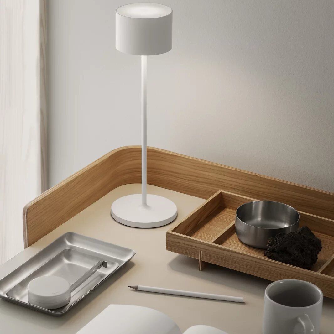 Mobile LED-Tischleuchte »FAROL« auf einem minimalistischen Schreibtisch mit Holzelementen. Auf dem Schreibtisch befinden sich eine offene Zeitschrift, eine Tasse, ein Tablett, ein Aschenbecher und ein Stift.